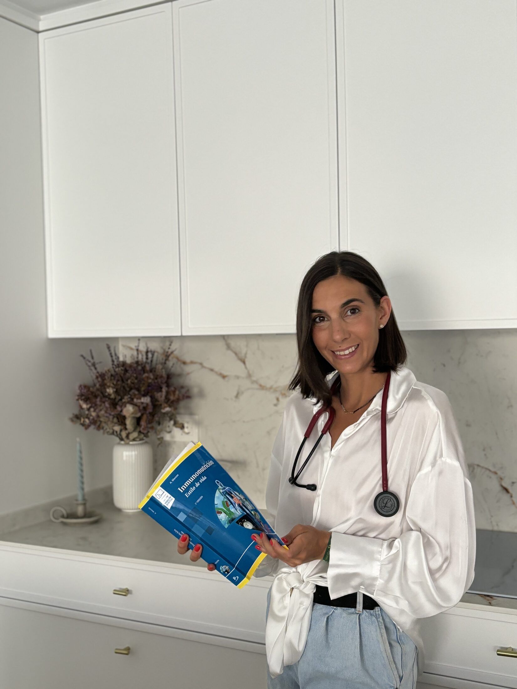 La doctora Isabel Sánchez Claros sonríe a cámara con un libro de inmunonutrición en las manos y el estetoscopio al cuello